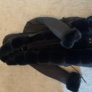 Svart pälsjacka med huva - Säljer en svart pälsjacka med fluffiga detaljer längs hela framsidan, huva och ärmslut. Jackan har dragkedja och en riktigt lyxig vibe, perfekt för kalla dagar när du vill sticka ut. Snygg och varm med en oversized känsla.