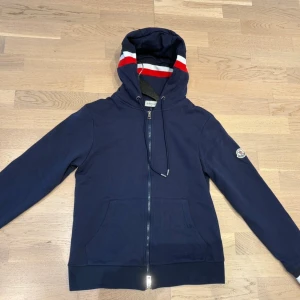 Marinblå Moncler hoodie med dragkedja - Snygg marinblå hoodie från Moncler med hel dragkedja, huva och fickor framtill. Ränder i rött, vitt och blått på huva och ärmslut ger en sportig vibe. Mjukt bomullsmaterial och klassisk Moncler-logga på ärmen. Tveka inte på att DM'a för frågor!