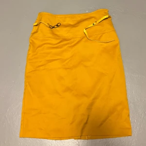 Gul kjol från DYA, storlek 38 - Gul kjol med coola detaljer som snörning och kedja i midjan. Kjolen har en rak, enkel form och är tillverkad i ett mjukt skönt material. Perfekt för dig som vill sticka ut med färg och stil.