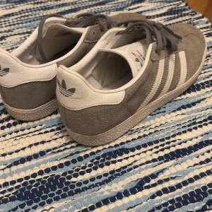 Grå Adidas Gazelle sneakers - Säljer ett par grå Adidas Gazelle sneakers med klassiska vita ränder och logga på hälen. Skorna har snörning, rund tå och platt sula. Ovandelen är i mjuk mocka och detaljer i vitt skinn. Perfekt för dig som gillar retrostil och vill ha bekväma sneakers.