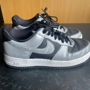 Nike Air Force 1 svart och silver - Nike Air Force 1 sneakers i svart och silver med lågt skaft. Ovandel i skinn med texturerade paneler och silvrig Swoosh med reptilmönster. Svarta snören och vit sula med AIR-detalj. Perfekt för dig som gillar klassisk streetstil.