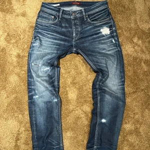 Blå slitna jeans från Jack & Jones - Säljer ett par blå jeans från Jack & Jones med snygga slitningar och tvättade detaljer. Modellen har klassisk femficksdesign, raka ben och normal passform. Jeansen är tillverkade i bomull och har en mörkblå ton med ljusare partier och slitna effekter på både fram- och baksida.