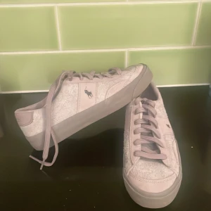 Ljusgrå sneakers från Polo Ralph Lauren - Snygga ljusgrå sneakers från Polo Ralph Lauren med klassisk logga på sidan. Skorna har rund tå, platt sula och snörning. Ovandelen är i en mix av mocka och syntet med en lite ruffad textur som ger extra karaktär. Perfekta för dig som gillar stilrena och sportiga skor.