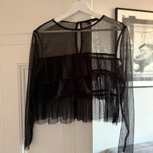 Svart transparent blus med volanger Zara - Supersnygg svart transparent blus från Zara med prickigt mönster. Blusen har långa ärmar, volangdetaljer framtill och knäppning i nacken. 
