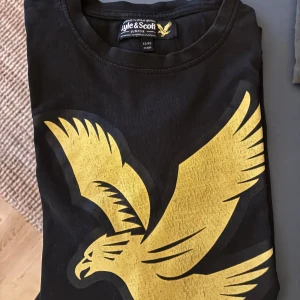 Svart t-shirt Lyle & Scott Junior - Svart t-shirt från Lyle & Scott Junior med stor gul fågel-logga på framsidan. Klassisk rund halsringning och korta ärmar. Perfekt för dig som gillar streetwear och vill sticka ut med en fet logga.