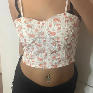 Blommig vit korsett-topp med smala band - Supersöt korsett-topp i vitt med blommigt mönster i rosa och grönt. Toppen har smala axelband och en croppad passform som framhäver midjan. Perfekt för sommarens alla tillfällen och lätt att matcha med både jeans och kjol.