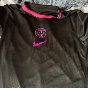 Svart PSG fotbollströja Nike S - Svart PSG fotbollströja från Nike med Dri-Fit material. Tröjan har korta ärmar, rosa detaljer vid kragen och ett rosa PSG-emblem samt Nike-logga med drip-effekt på bröstet. Snyggt mönster i tyget och trycket 'G10' på ryggen. Perfekt för dig som gillar streetstyle och fotboll.