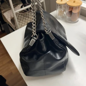 Svart axelväska med kedjerem - Säljer en svart axelväska i skinn med coola silverfärgade kedjeremmar. Väskan har en mjuk, lite boxig form och stilrena sömmar som ger en modern vibe. Perfekt för dig som vill ha en snygg och praktisk väska med attityd.