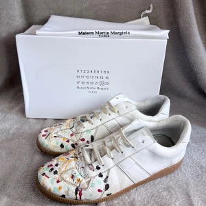 Maison Margiela vita sneakers med färgstänk - Maison Margiela sneakers i vitt skinn med coola färgstänk i rött, gult, svart och grönt på tån. Klassisk låg modell med snörning och gummisula i beige. Snygg och unik design som sticker ut, perfekt för dig som gillar statement pieces.