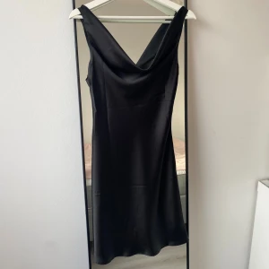 Svart satinklänning från Even&Odd - Supersnygg svart klänning i satin från Even&Odd. Klänningen har en elegant draperad v-ringning och är ärmlös med axelband. Den har en slät, glansig yta och en klassisk, figurnära passform som faller snyggt längs kroppen. Det förekommer något slitage i form av revor 