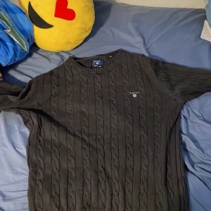 Svart kabelstickad tröja från GANT - Svart kabelstickad tröja från GANT i premium bomull. Klassisk rund halsringning och broderad logga på bröstet. Perfekt för dig som gillar stilren och tidlös design med en preppy touch.