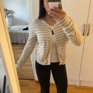 Beige och vit randig kofta från H&M - Säljer en beige och vit randig kofta från H&M i storlek M. Koftan har rund hals, långa ärmar och knappar framtill. Mjuk och tunn stickad känsla, perfekt för lager-på-lager. Står M men passar perfekt till en person som också har S