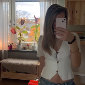 Vit croppad blus med v-ringning - Säljer en vit blus med korta ärmar och snygg v-ringning. Toppen är croppad och har tre dekorativa knappar framtill. Passformen är normal. Stilrena och trendiga.