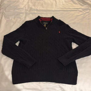 Mörkblå stickad tröja Polo Ralph Lauren Half Zip - Mörkblå kabelstickad tröja. Polo Ralph Lauren med halv dragkedja och röd broderad logga på bröstet. Storlek M
