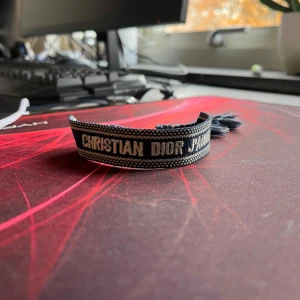 Blått Christian Dior J'Adior armband - Snyggt blått armband från Christian Dior med texten 'J'Adior' broderad i vitt. Armbandet har en vävd design med vita detaljer och avslutas med stiliga blå och vita tofsar. Perfekt accessoar för att lyfta din outfit med lite lyxig vibe.