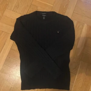 Svart kabelstickad tröja från GANT - Svart kabelstickad tröja från GANT i stretchig bomull. Klassisk rund halsringning och diskret logga på bröstet. Långärmad modell med ribbade muddar och smal passform. Perfekt för lager-på-lager under höst och vinter.