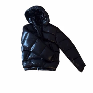 Svart pufferjacka från Zara - Svart pufferjacka från Zara med glansig finish och huva. Jackan har breda, quiltade sektioner och dragkedja framtill. Perfekt för kalla dagar och har en modern, loose passform som ger en cool vibe. Materialet är syntet och jackan är långärmad. Jackan är använd knappt 3 gånger! Helt Ny!