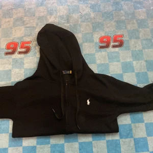 Svart hoodie från Polo Ralph Lauren - Svart hoodie från Polo Ralph Lauren med klassisk vit broderad logga på bröstet. Tröjan har dragkedja framtill, huva med snörning och känguruficka. Perfekt för en chill och stilren look.