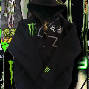 Monster x Fox svart hoodie XL - Svart hoodie från Fox och Monster Energy med gröna och vita broderade loggor, dragkedja och huva. Snygga detaljer med stora tryck och brodyr på bröstet och ärmen samt nummer 4 på huvan. Perfekt för dig som gillar streetwear och motorsport.