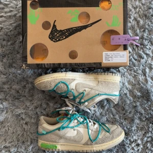 Nike Dunk Low Off-White Lot 36/50 - Nike Dunk Low Off-White Lot 12/50 med beige och grå mocka, vita detaljer och turkosa snören. Snygga Off-White-tag och textdetaljer på sidan. Unik design med extra snörning och klassisk Dunk-sula. Kommer med originalkartong och Off-White-påse.