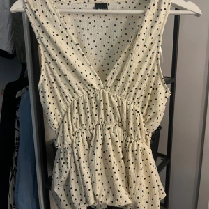 Prickig ärmlös topp från Gina Tricot - Supersöt ärmlös topp från Gina Tricot i beige med svarta prickar. Toppen har v-ringning, volangdetaljer och en luftig, rynkad passform som ger en avslappnad vibe. Perfekt för varma dagar och enkel att styla med jeans eller kjol.