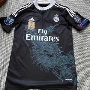 Real Madrid Ronaldo 7 svart fotbollströja - Säljer en svart Real Madrid fotbollströja från Adidas med Ronaldo 7 på ryggen. Tröjan har ett unikt drakmotiv framtill, vita detaljer och märken som FIFA World Champions 2014 och Champions League-patchar på ärmarna. Materialet är lätt och andas, Climacool-teknologi.