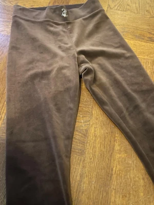 Bruna velourbyxor Juicy Couture XXS - Säljer ett par bruna velourbyxor från Juicy Couture i storlek XXS. Byxorna har bootcut-ben och låg midja, med glittriga strasstenar som bildar texten 'Juicy' på baksidan. Mjukt material och klassisk Juicy-stil, perfekt för dig som gillar Y2K-vibe.