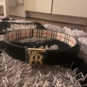 Burberry bälte med guldspänne - Säljer ett snyggt Burberry bälte med klassiskt rutigt mönster i beige, svart och rött på ena sidan och svart skinn på andra. Spännet är i guld med Burberrys ikoniska TB-logga. Perfekt accessoar för att lyfta din outfit.