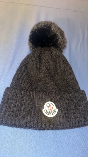 Svart Moncler mössa med pälsboll - Svart ribbstickad mössa från Moncler med stor fluffig pälsboll på toppen. Klassisk uppvikt kant och Moncler-logga framtill. Perfekt accessoar för kalla dagar med exklusiv känsla och snygg design.