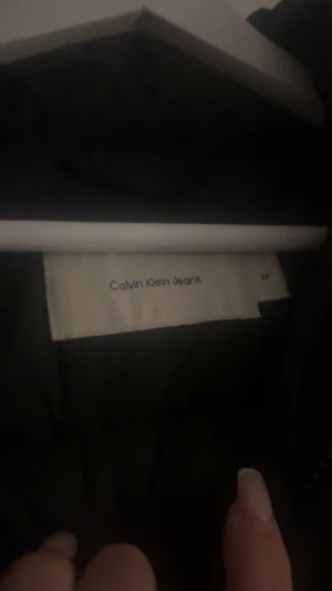 Svart pufferjacka Calvin Klein Jeans - Svart pufferjacka från Calvin Klein Jeans med klassisk CK-logga på bröstet. Jackan har hög krage, långa ärmar och dragkedja framtill. Själv brukar jag ha S i tröjor och denna jacka passar perfekt för mig. 