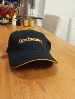 Svart Continental keps med gul brodyr - Snygg svart keps från Continental med gul broderad logga framtill och gul kant på skärmen. Klassisk form med böjd skärm och justerbar passform. Perfekt för dig som gillar streetstyle eller vill sticka ut med en cool accessoar.