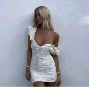 Vit kort klänning med volangdetaljer - Supersöt vit kort klänning med volanger på axel och ärm, snörning framtill och figurnära passform. Perfekt för sommarens alla tillfällen och ger en romantisk vibe. Klänningen har en axelbandslös look på ena sidan och är tillverkad i ett lätt material. Storlek S men passar xs-s. Har bara lite defekter i innertyget som jag sytt igen men synd inte när man har på sig den❤️