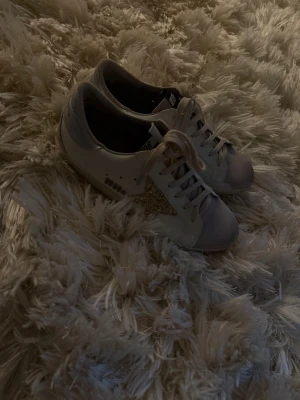 Golden Goose  - Golden Goose sneakers i vitt skinn med beige mockadetaljer och snörning. Sidan pryds av en glittrig guldstjärna som ger extra edge. Baksidan har silvriga detaljer med logga. Platt sula och rund tå, perfekt för en trendig streetstyle.