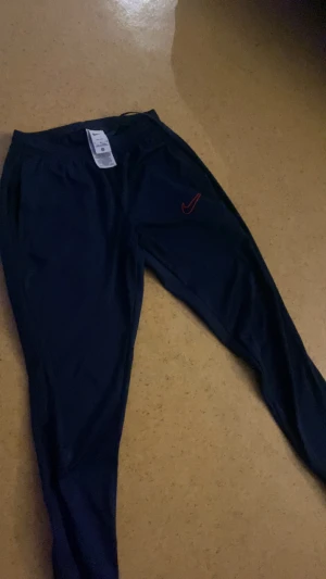 Blå Nike träningsbyxor M - Snygga blå träningsbyxor från Nike i storlek M. Byxorna har ett rött Nike Swoosh broderat på benet och är gjorda i ett lätt och stretchigt syntetmaterial. De har smal passform och elastisk midja med snörning för extra komfort.