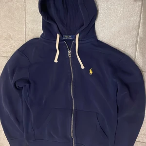 Marinblå hoodie från Polo Ralph Lauren - Snygg marinblå hoodie från Polo Ralph Lauren med gul broderad logga på bröstet. Tröjan har vit snörning i huvan, dragkedja framtill och två fickor. Tillverkad i mjuk bomull och har en klassisk, normal passform. Perfekt för chill dagar och streetwear. Är i storlek M men sitter mer som S