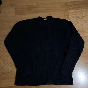 Svart kabelstickad tröja - Kabelstickad mörkblå tröja från Lindex med rund hals och långa ärmar. Tröjan har ett klassiskt mönster med vertikala flätor och ribbade muddar vid ärmslut och nederkant. Perfekt för dig som gillar enkel och stilren design.