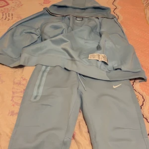 Nike ljusblå mjukisbyxor med snörning - Snygga ljusblå mjukisbyxor från Nike med klassisk logga på benet. Byxorna har snörning i midjan och en sportig look med diskreta ränder längs fickorna. Perfekt för chill eller träning, riktigt sköna och stilrena.