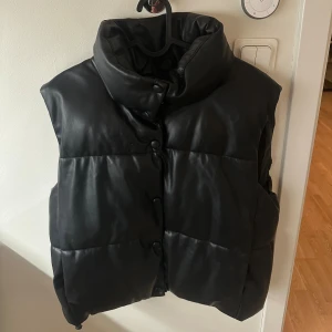 Svart dunväst från Zara - Svart dunväst från Zara med hög krage, skinlinande yta och tryckknappar framtill. Skulle säga att den passar alla storlekar mellan XS-L.