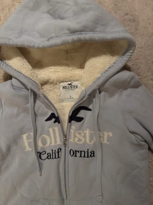 Ljusblå hoodiejacka från Hollister - Mysig ljusblå hoodiejacka från Hollister med broderad logga och text på bröstet. Jackan har dragkedja, snörning i huvan och är fodrad med fluffigt, krämfärgat teddyfoder. Perfekt för kyliga dagar och riktigt skön att ha på sig.