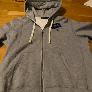 Grå hoodie från Polo Ralph Lauren - Snygg grå hoodie från Polo Ralph Lauren med dragkedja och vit snörning i huvan. Storlek S men passar större (någon som är ungefär 182-187cm lång). priset kan diskuteras