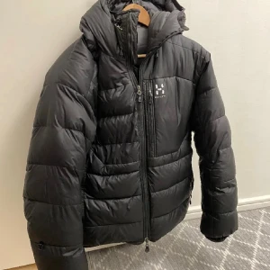 Svart dunjacka från Haglöfs - Svart dunjacka från Haglöfs med puffig design och huva. Jackan har dragkedja framtill, två sidofickor med dragkedja och logga på bröstet. Perfekt för kalla dagar och riktigt skön att bära. Klassisk och stilren modell.