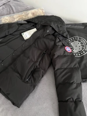 Canada Goose jacka  - Säljer en svart dunjacka från Canada Goose med huva och avtagbar pälskant. Jackan har klassisk logga på ärmen, dragkedja och knappar framtill samt stora fickor. Perfekt för kalla vinterdagar och riktigt snygg streetstil.