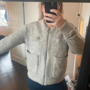 Grå ulljacka med dragkedja - Mysig grå jacka i ull med rund halsringning och dragkedja framtill. Jackan har två stora fickor nertill och två mindre bröstfickor, 