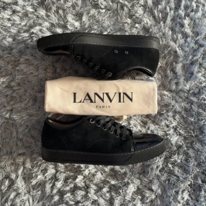 Svarta sneakers från Lanvin - Säljer ett par svarta Lanvin sneakers med snygg platt sula och rund tå. Ovandelen är i mocka med detaljer i blankt skinn vid tån, och de har klassisk snörning. Insidan är fodrad i beige skinn för extra komfort. Perfekt för dig som gillar stilrena och lyxiga sneakers.