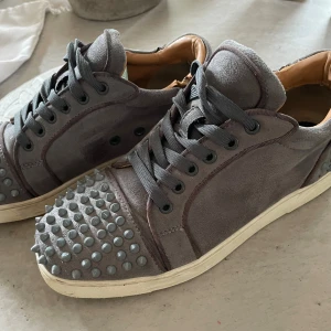 Grå Christian Louboutin sneakers med nitar - Säljer ett par grå sneakers från Christian Louboutin med ikoniska röda sulor. Skorna är i mocka och har coola nitar på tå och häl, samt svarta snören. Insidan är i beige skinn och de har en platt sula. Perfekt för dig som vill sticka ut med din stil. Passar 40 till  41,5