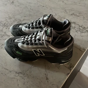 New Balance 1000 svarta sneakers - Säljer ett par New Balance 1000 sneakers i svart och silver med gröna detaljer. Skorna har mesh och syntetmaterial, snörning och en chunky platt sula. Ikonisk N-logga på sidan och 1000-märkning. Perfekta för dig som gillar sportig och modern stil.