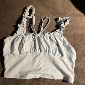 Ljusblå croppad linnetopp med volangband - Supersöt ljusblå croppad linnetopp med volangprydda axelband och smala knytband i ryggen. Toppen har en lätt rynkad detalj framtill och är tillverkad i mjukt bomullsmaterial. Perfekt för varma dagar och en chill stil.