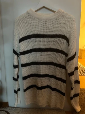 Vit och svart randig stickad tröja - Säljer en vit stickad tröja med svarta breda ränder. Tröjan har en rundad hals och långa ärmar, perfekt för dig som gillar en chill och klassisk stil. Materialet är grovstickat och ger en skön och avslappnad känsla.