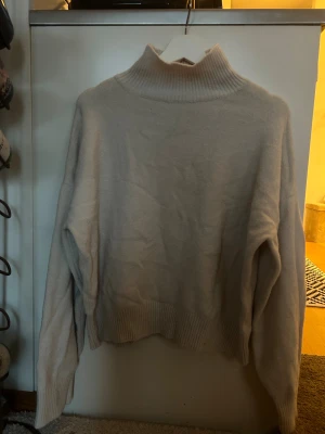 Vit polotröja med ribbad krage - Säljer en vit polotröja med bred, ribbad krage och muddar. Tröjan har en loose passform och är långärmad, perfekt för kyliga dagar. Materialet känns mjukt och mysigt, och färgen är neutral vilket gör den enkel att matcha med andra plagg. (ljuset gör färgen orättvis)