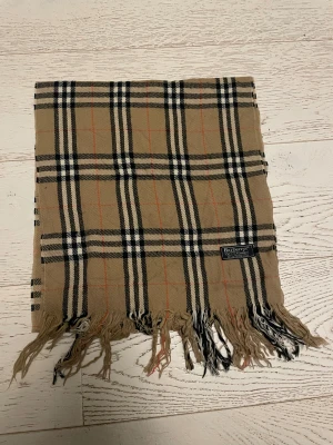 Rutig beige halsduk från Burberrys - Snygg klassisk halsduk från Burberrys i 100% kashmir. Beige botten med rutigt mönster i svart, vitt och tunna röda linjer. Mjukt material och fransar i ändarna. Perfekt för dig som vill ha en ikonisk accessoar med lyxig känsla.